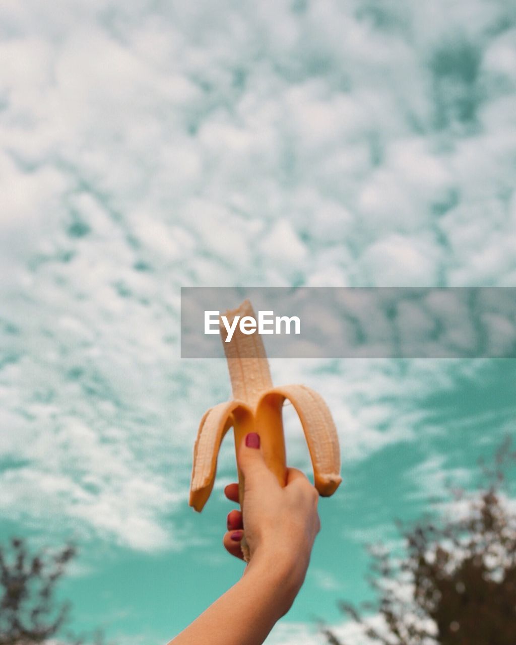 Cropped hand of woman having banana against cloudy sky