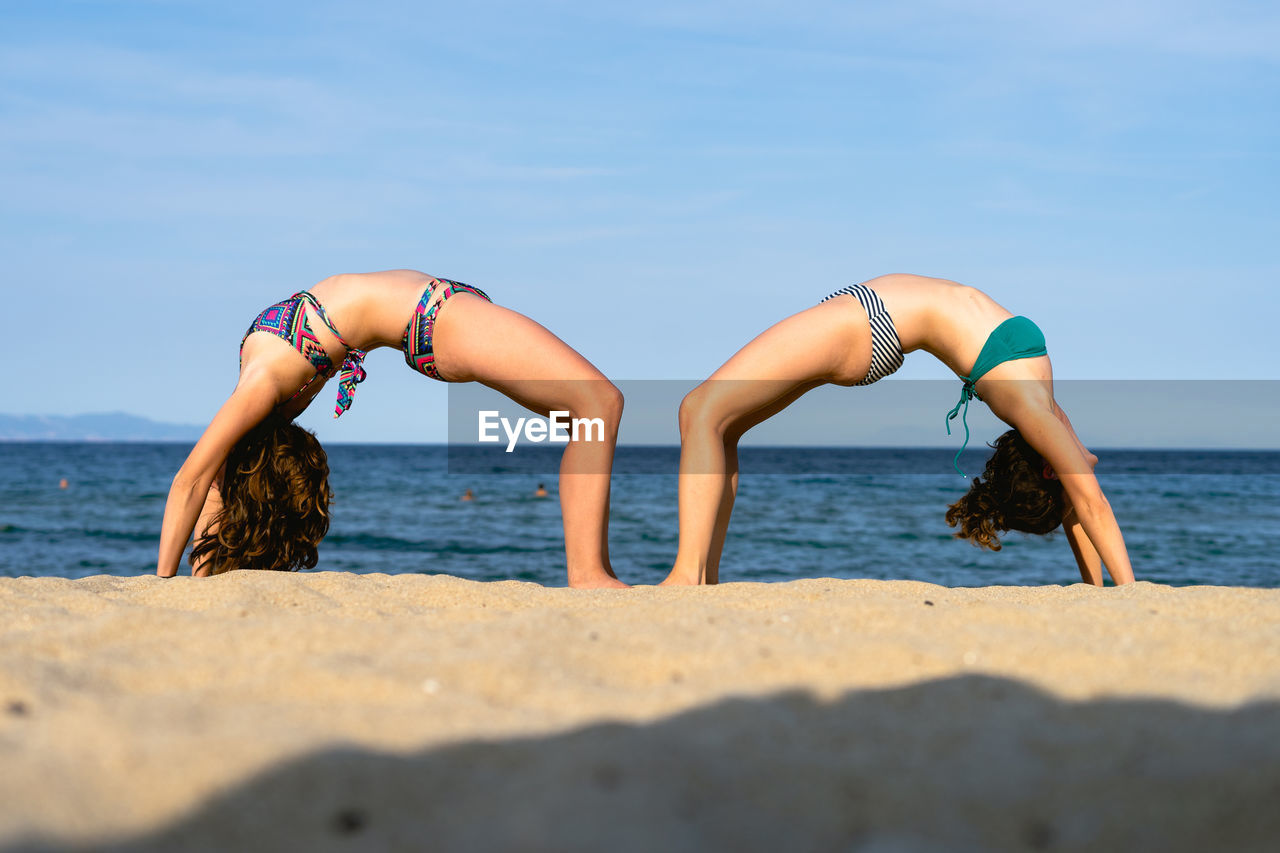 Side view of friends wearing bikinis exercising at beach against sky