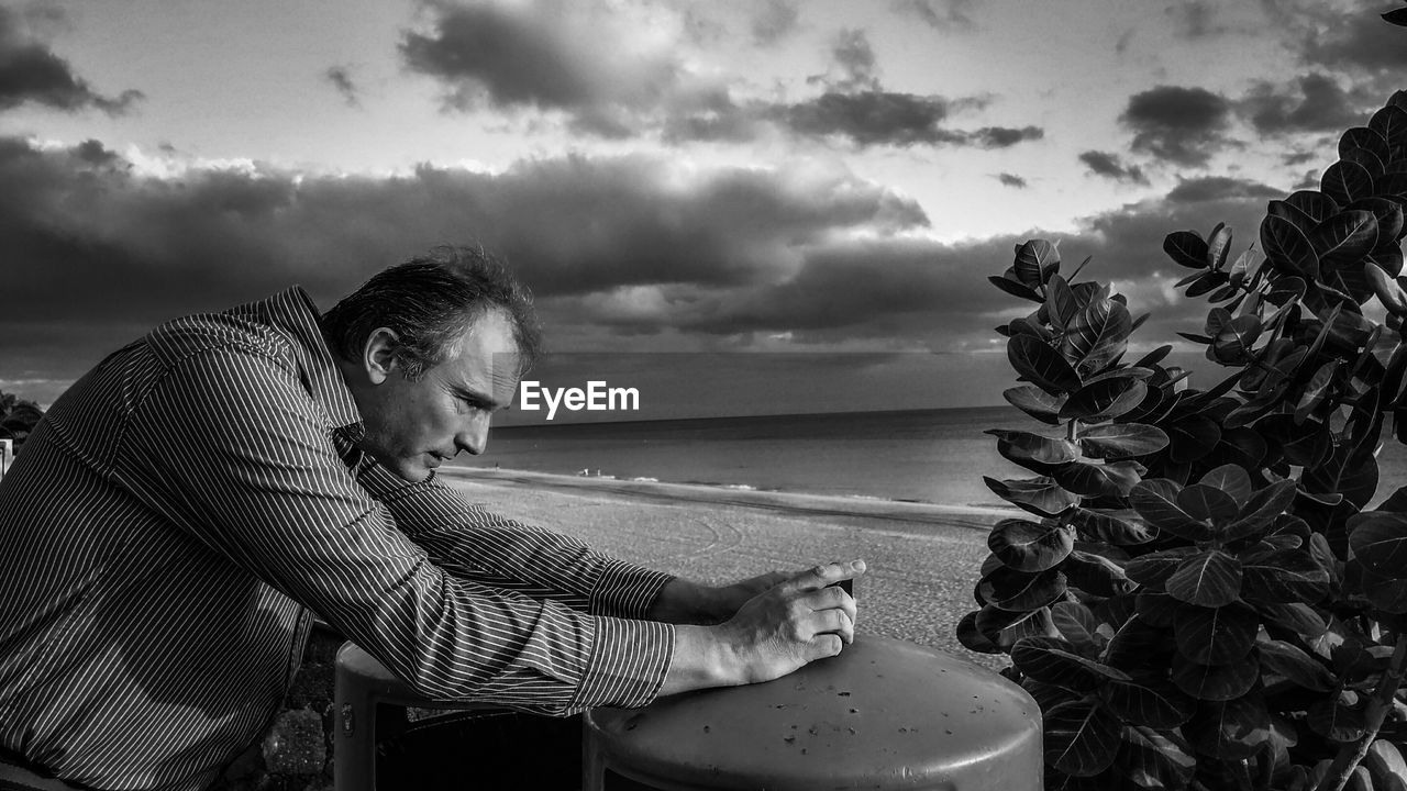 Side view of man photographing plant at beach against sky