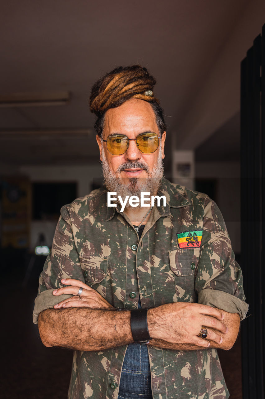 Bearded mature male in casual outfit with sunglasses and dreadlocks standing in light apartment and looking at camera