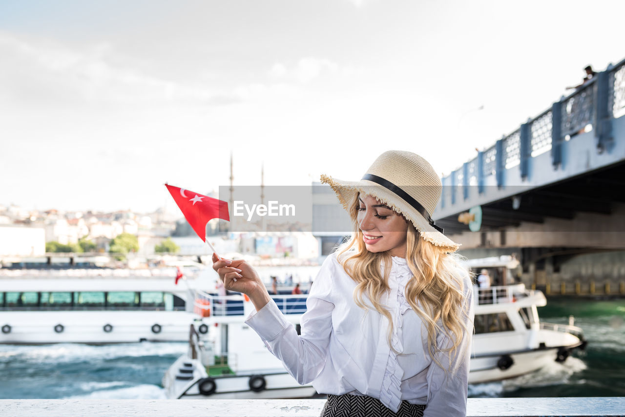 Young woman in hat holding flag while standing in boat against sky