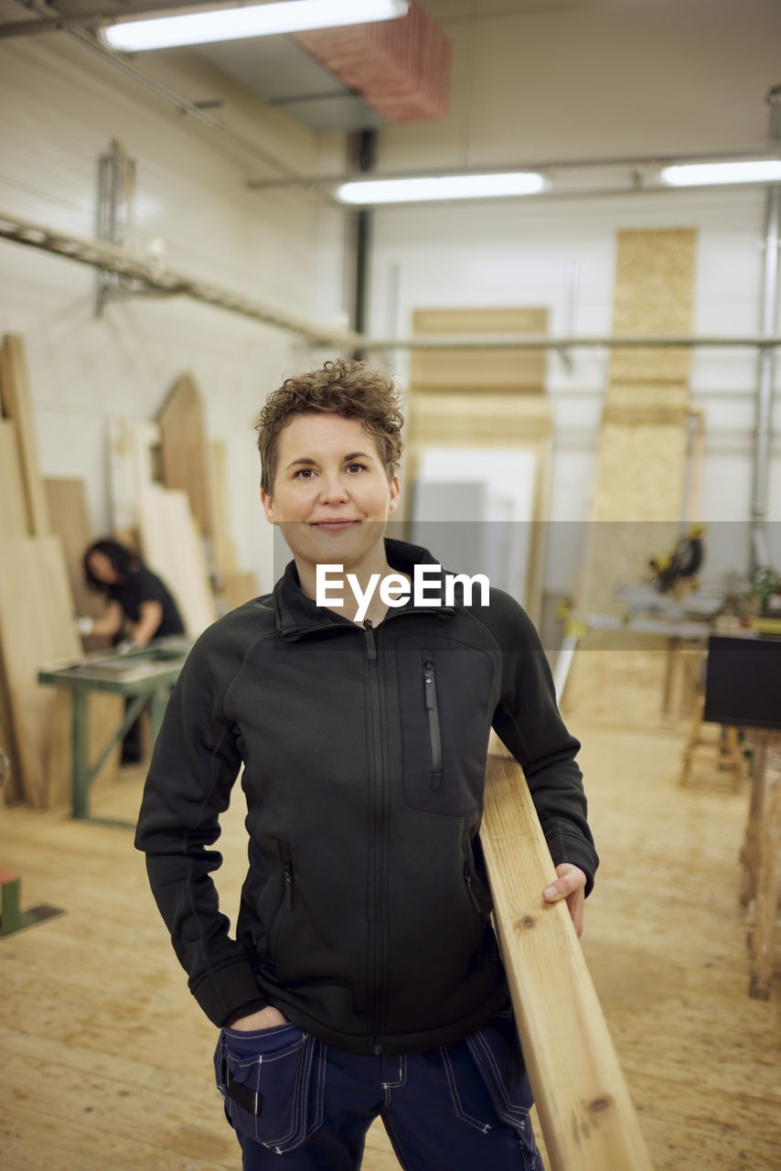 Portrait of confident mature female carpenter carrying plank while standing in workshop