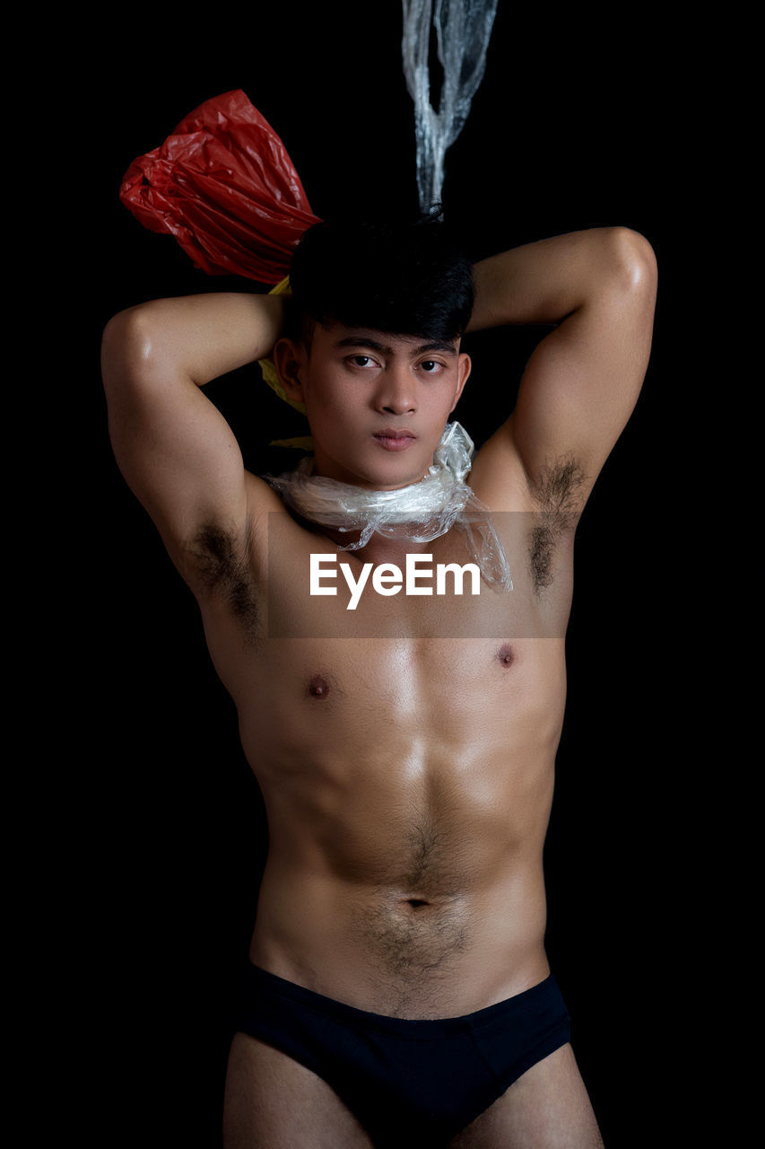 Portrait of shirtless young man with plastic around neck against black background