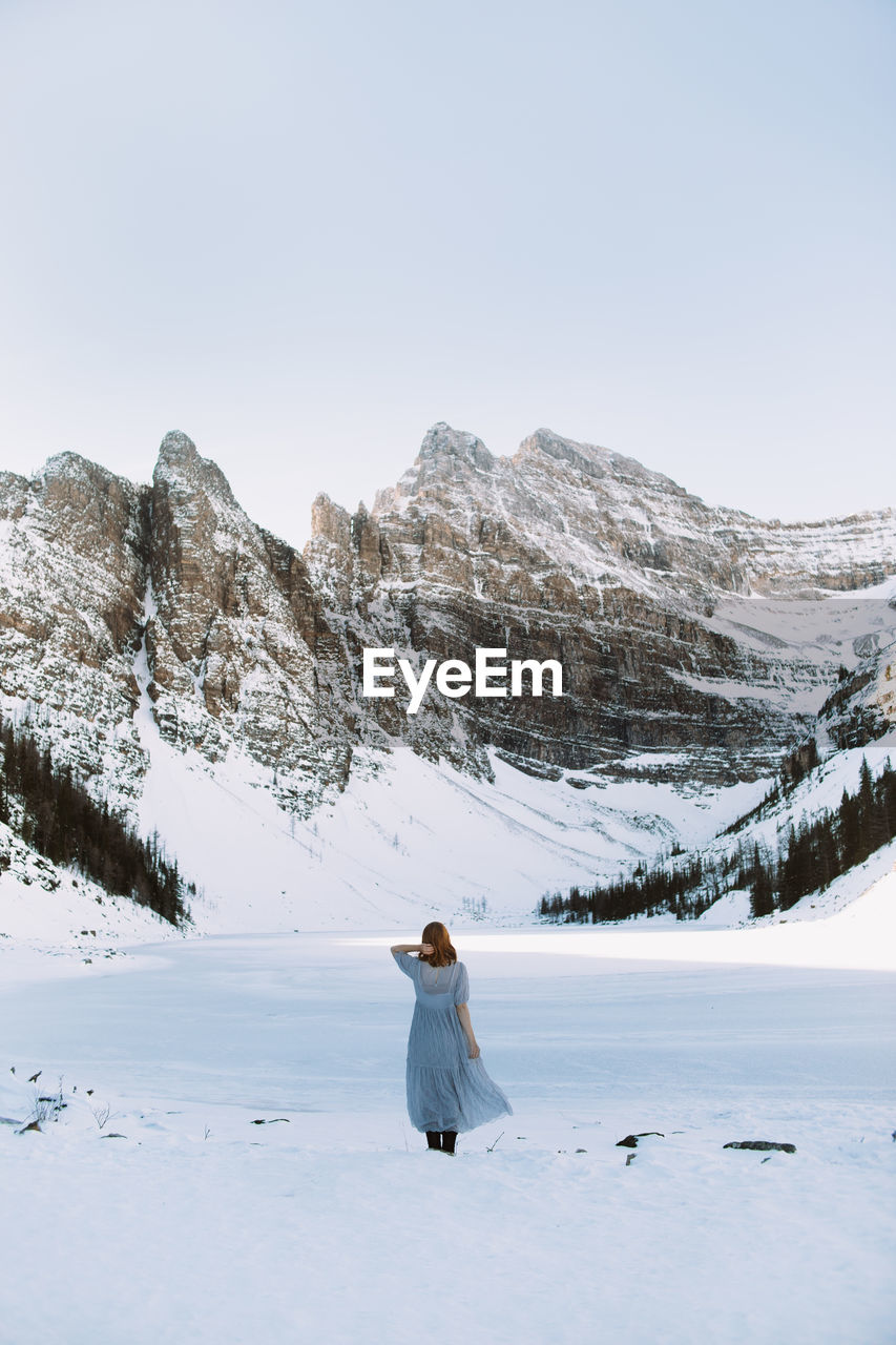 Back view of anonymous woman in dress touching hair and standing near frozen lake louise against snowy mountain range on winter day in alberta, canada