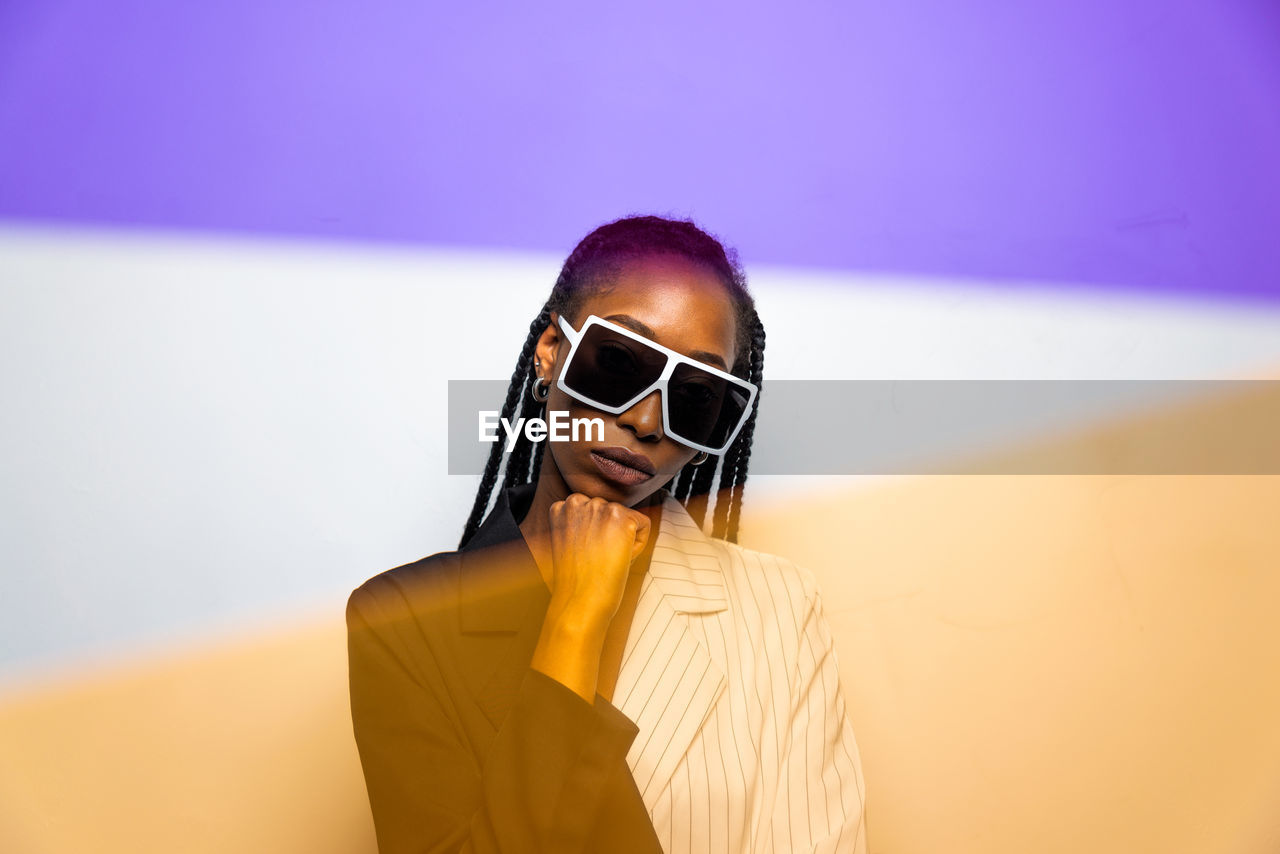 Portrait of young woman wearing sunglasses standing against wall