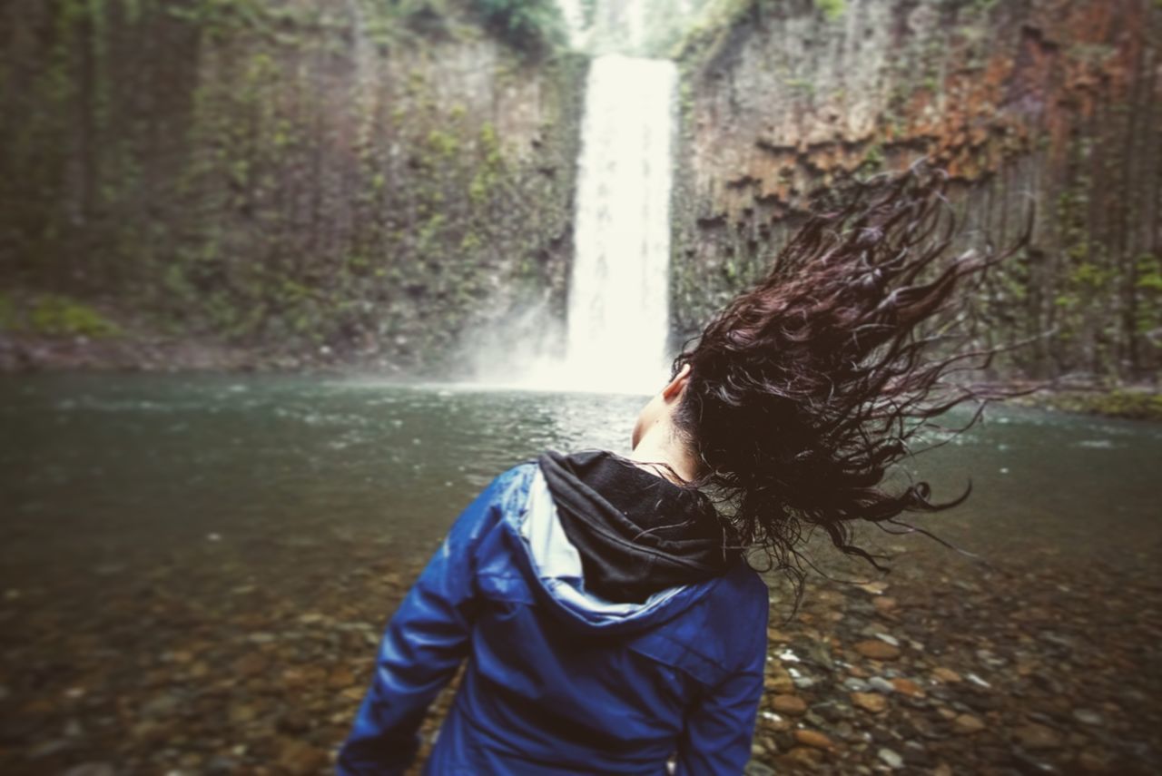 Rear view of woman shaking head while standing against waterfall