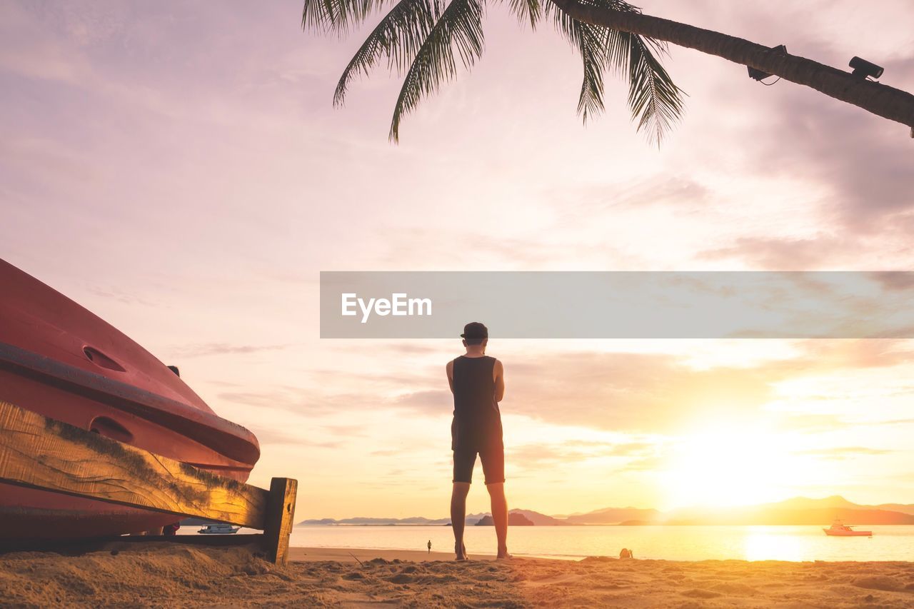 Rear view of man standing at beach against sky during sunset