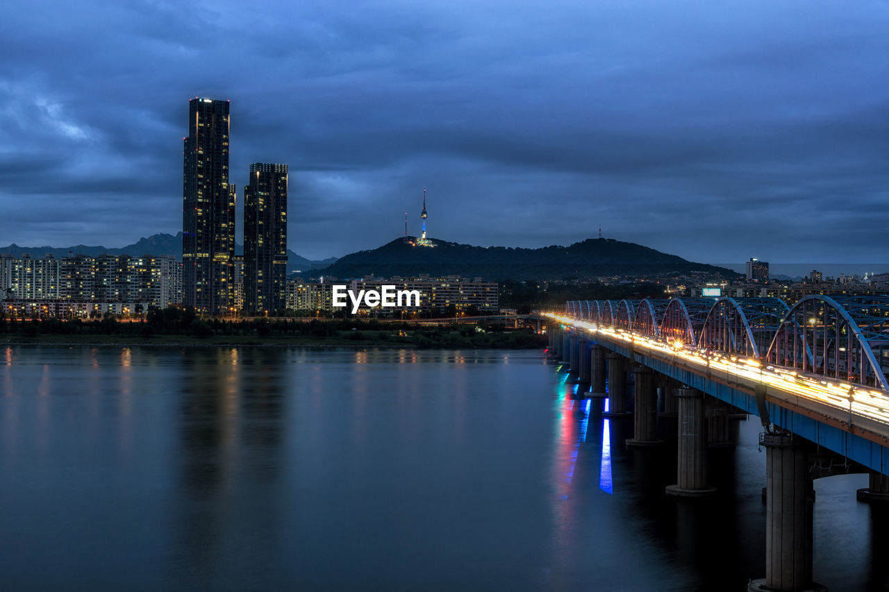 Illuminated bridge over river by buildings against sky at dusk