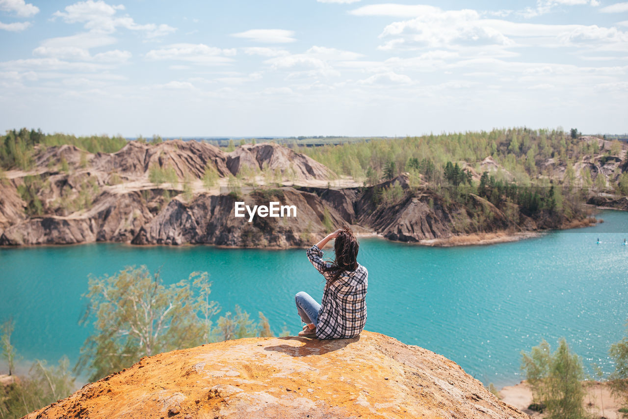 rear view of woman sitting on rock by lake against mountain