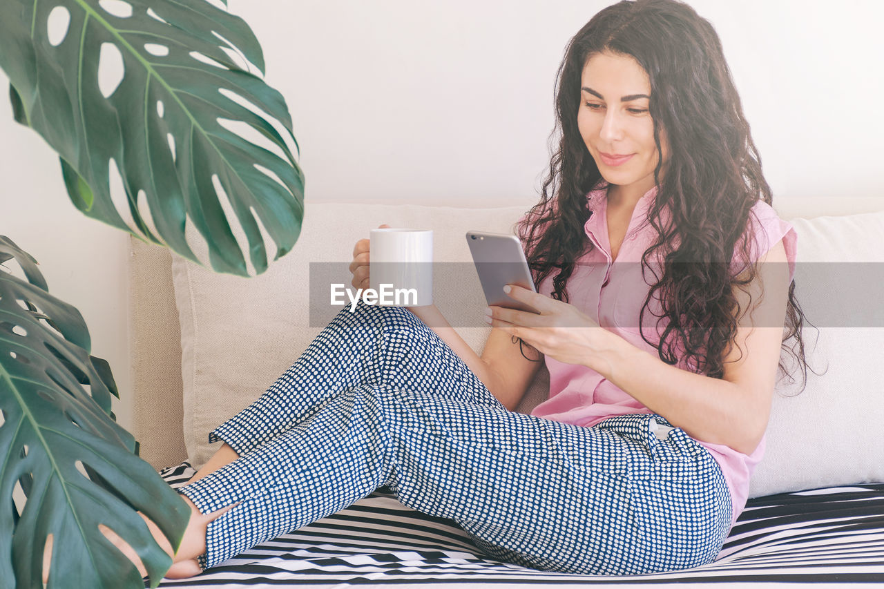 Relaxing woman spending time in the morning using mobile phone and drinking coffee on sofa at home.