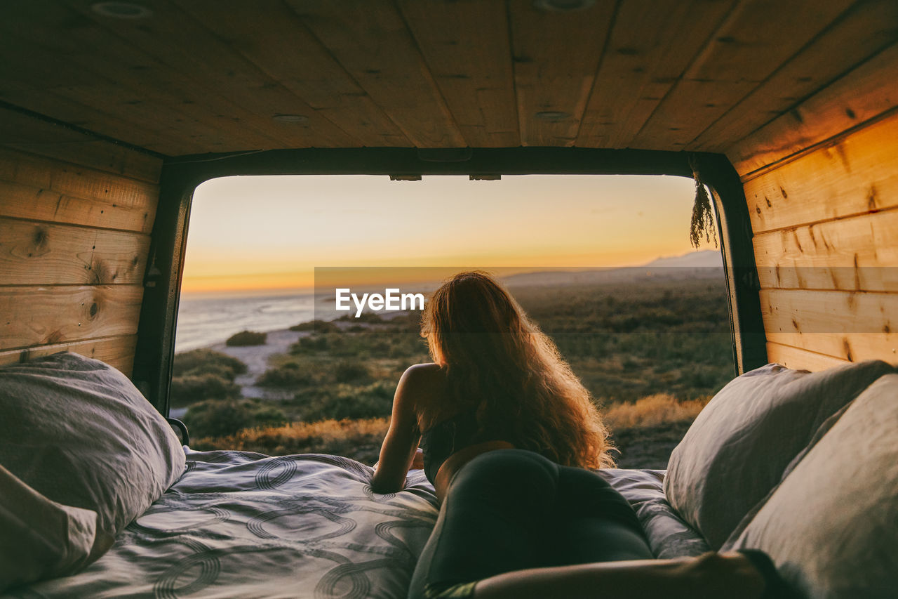 Young woman looking out to ocean from bed of camper van in mexico.