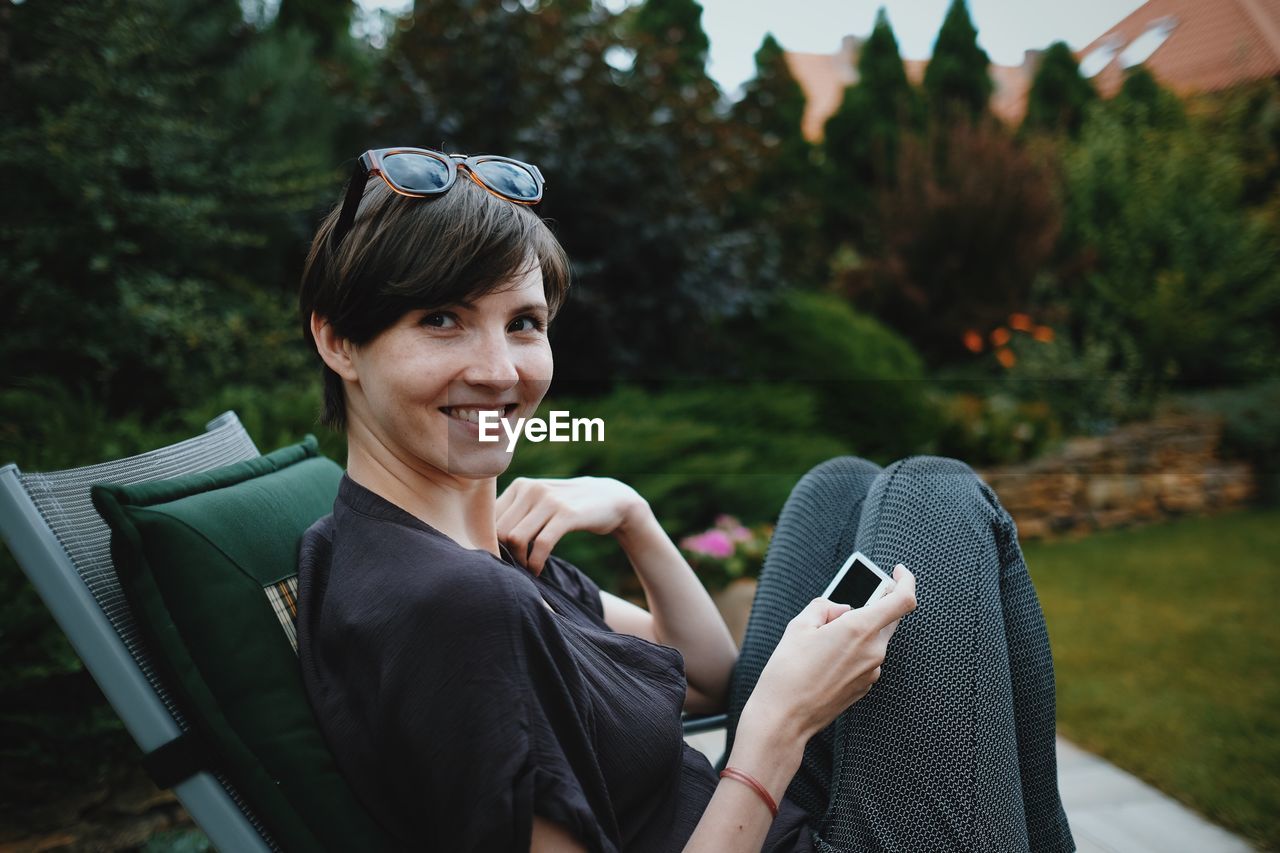 PORTRAIT OF SMILING YOUNG WOMAN USING SMART PHONE WHILE SITTING ON CAMERA