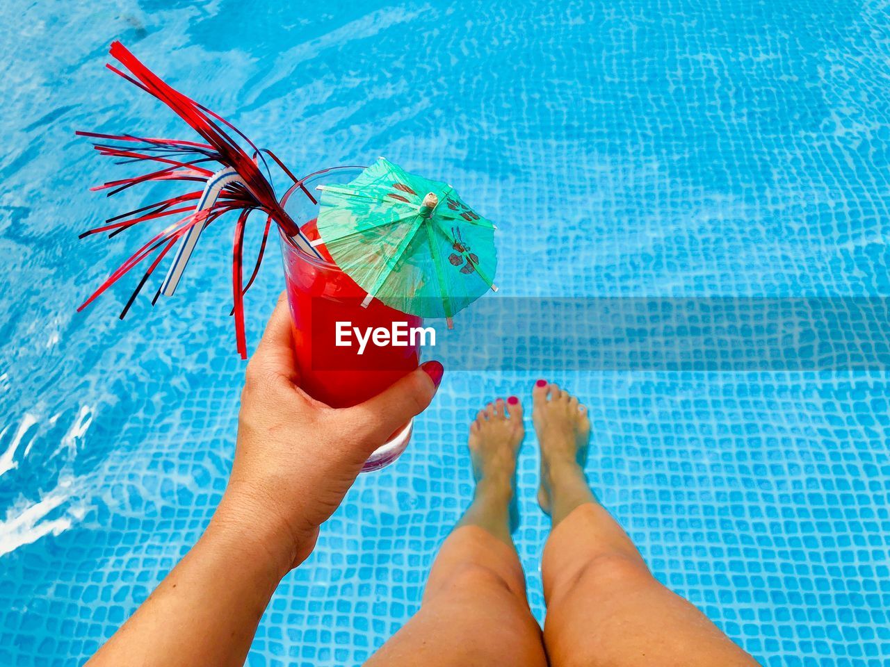 Low section of woman holding drink while sitting by swimming pool