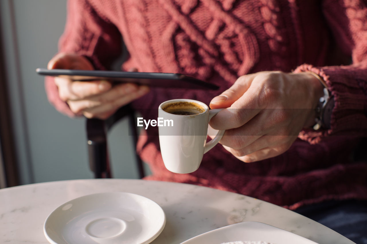 Midsection of man holding coffee cup using digital tablet in cafe