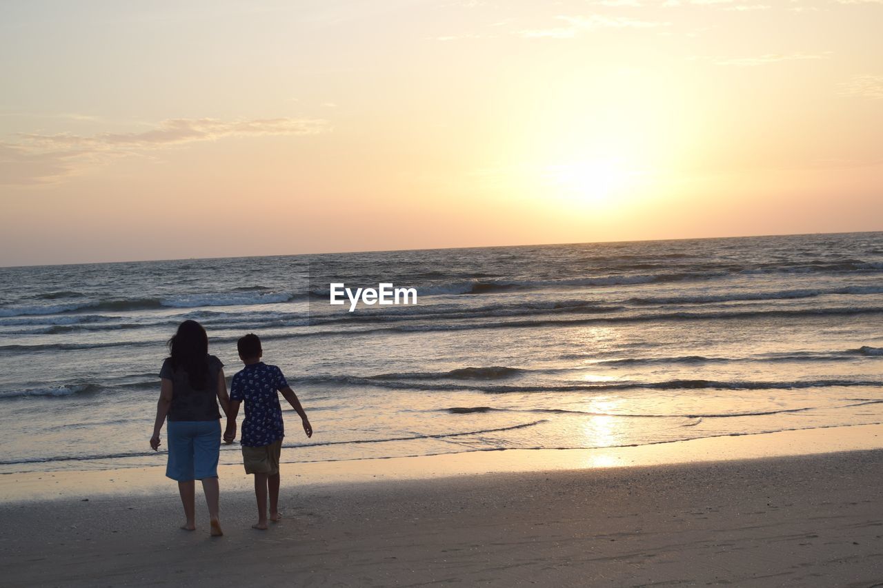 REAR VIEW OF WOMEN STANDING AT BEACH AGAINST SKY DURING SUNSET