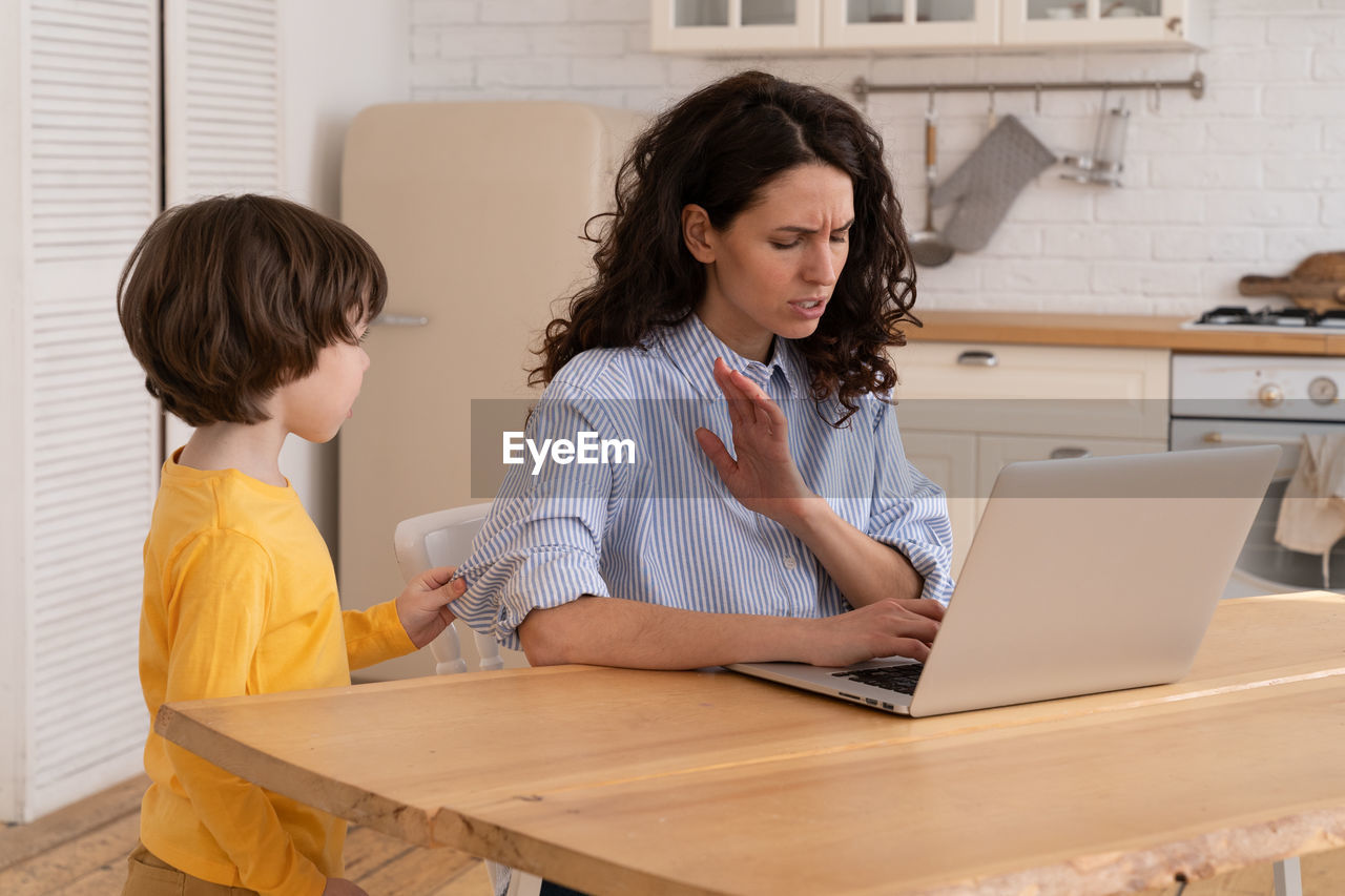 Mom sits by table at home office during lockdown, working on laptop, kid distracts and making noise