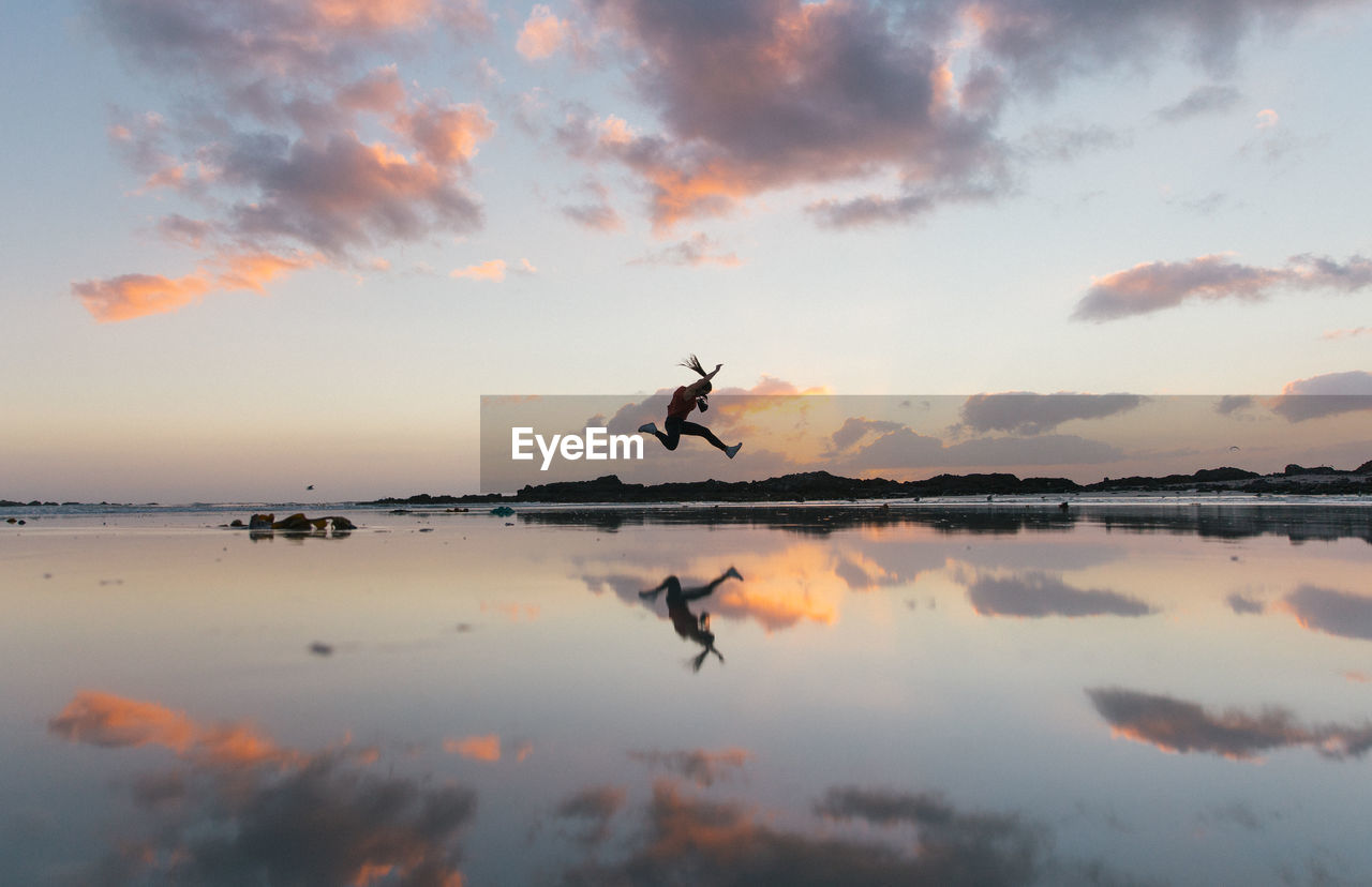 Woman jumping over lakeshore against sky during sunset