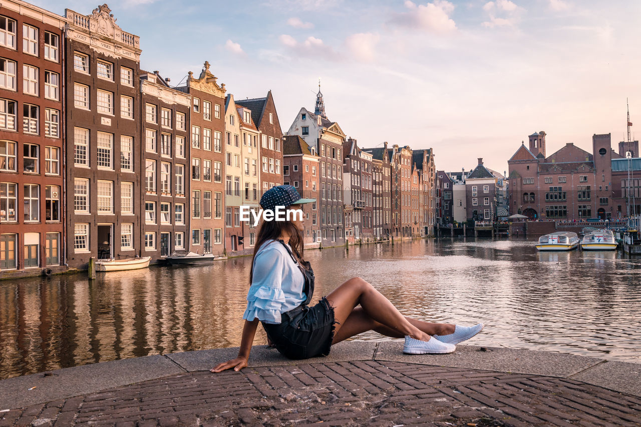 Side view of woman sitting by canal against buildings in city