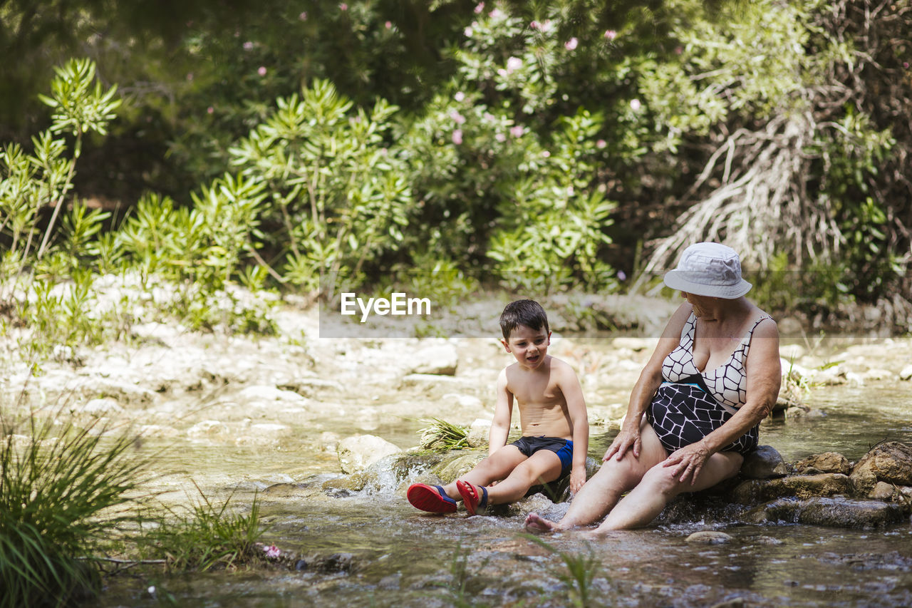 Grandmother and grandson talking while sitting on stone in river at forest