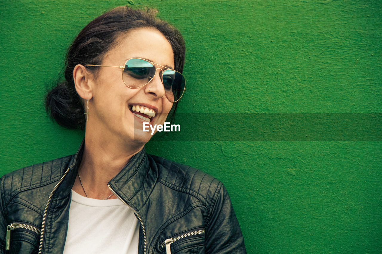 PORTRAIT OF SMILING YOUNG MAN WEARING SUNGLASSES AGAINST WALL