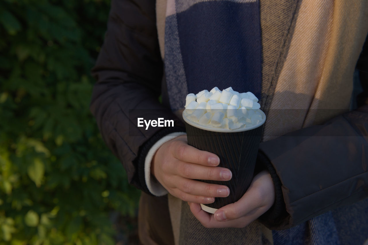 Warm evening sun highlights marshmallows atop of hot takeaway cacao drink. held by a young women