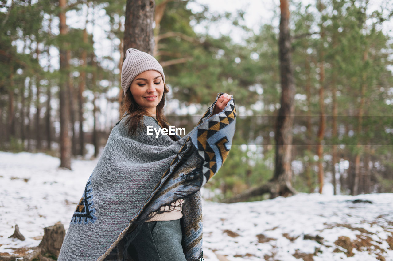 Portrait of young brunette beautiful woman in hat and grey poncho in winter forest