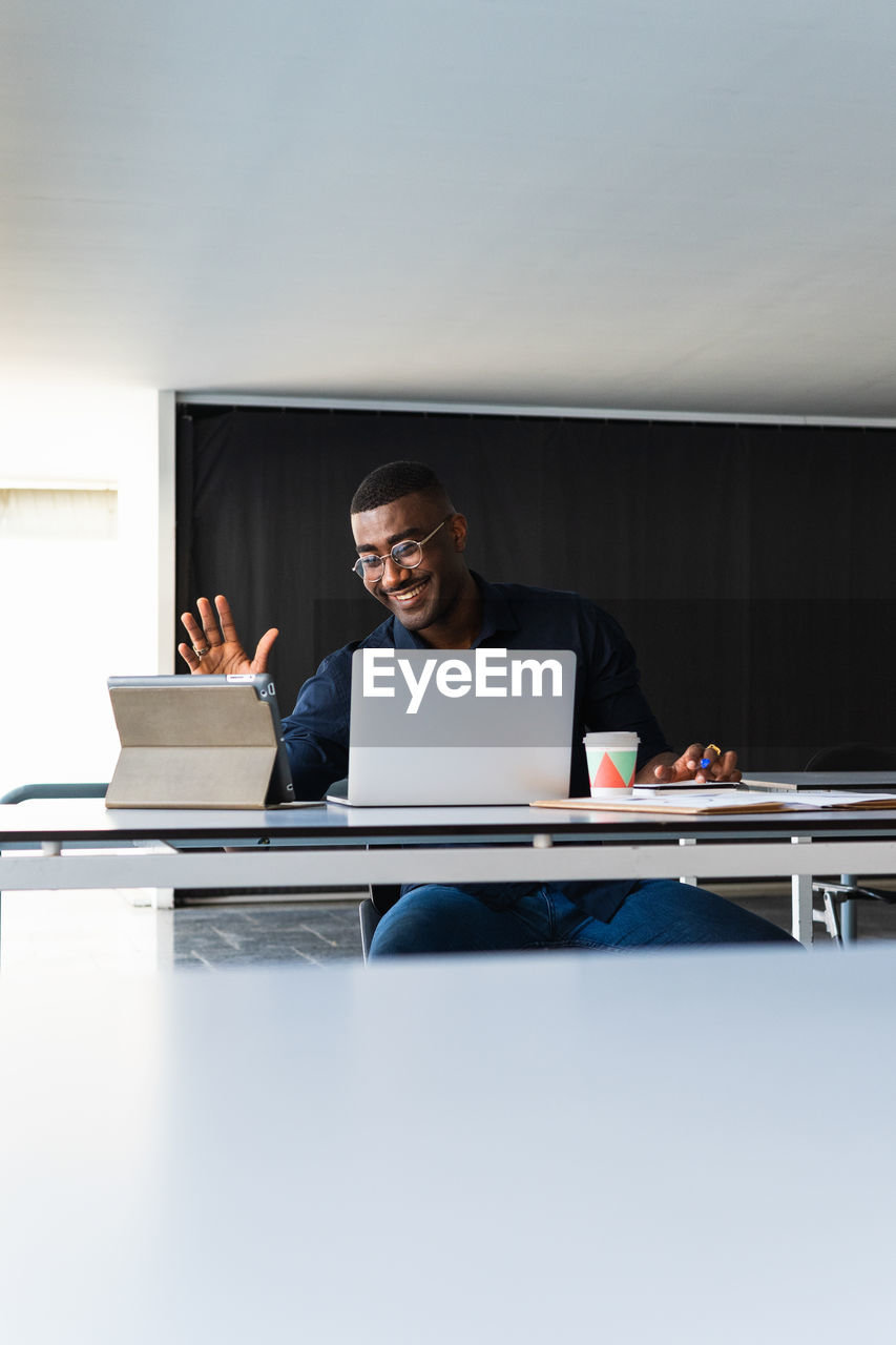Positive african american adult male in formal clothes and eyeglasses sitting at table while having video call on tablet near netbook and documents with takeaway drink in bright office