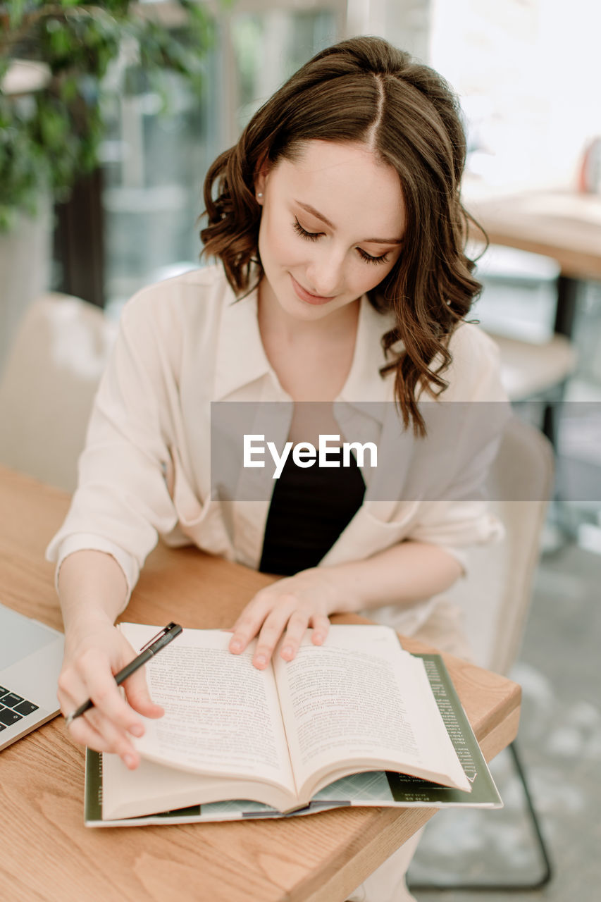 Young dark-haired woman sitting at the table with a laptop and book smiling