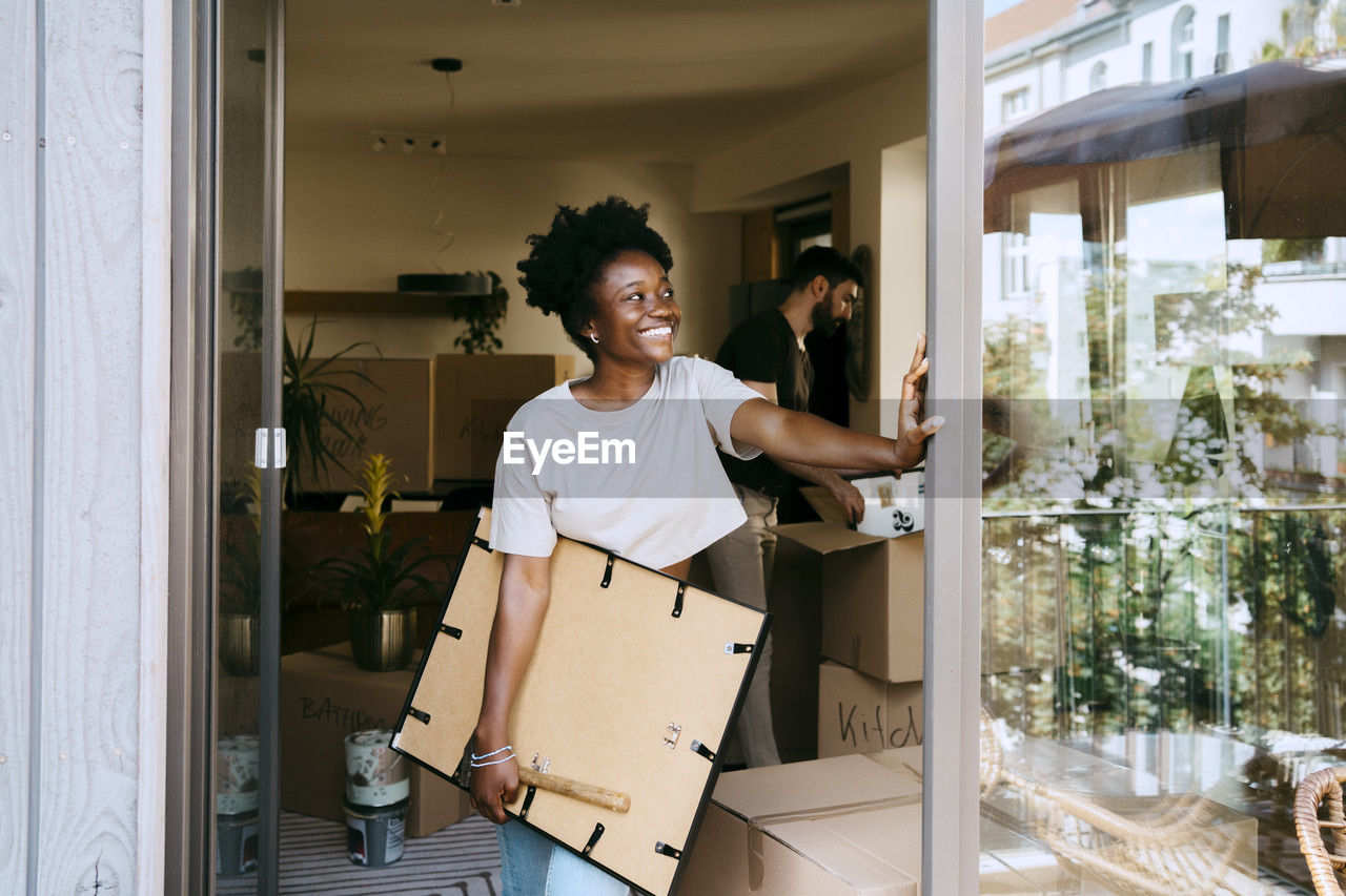 Smiling woman looking away holding frame while leaning on wall at new home