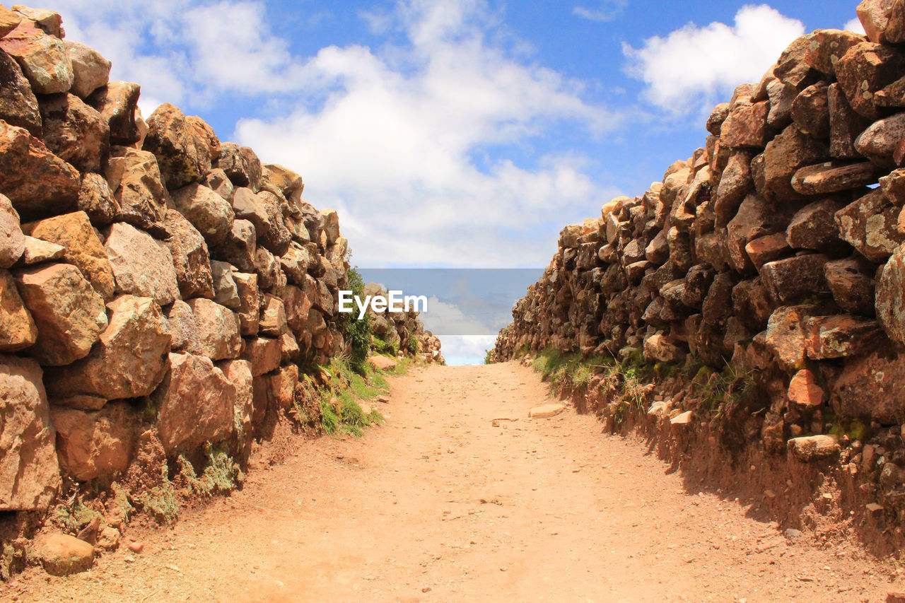 Empty footpath amidst stone wall against sky