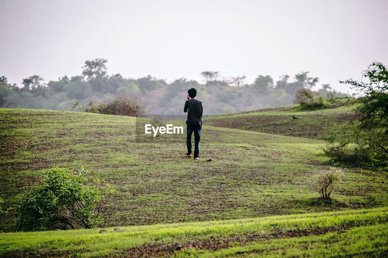 Rear view of young man walking on grassy hill against clear sky