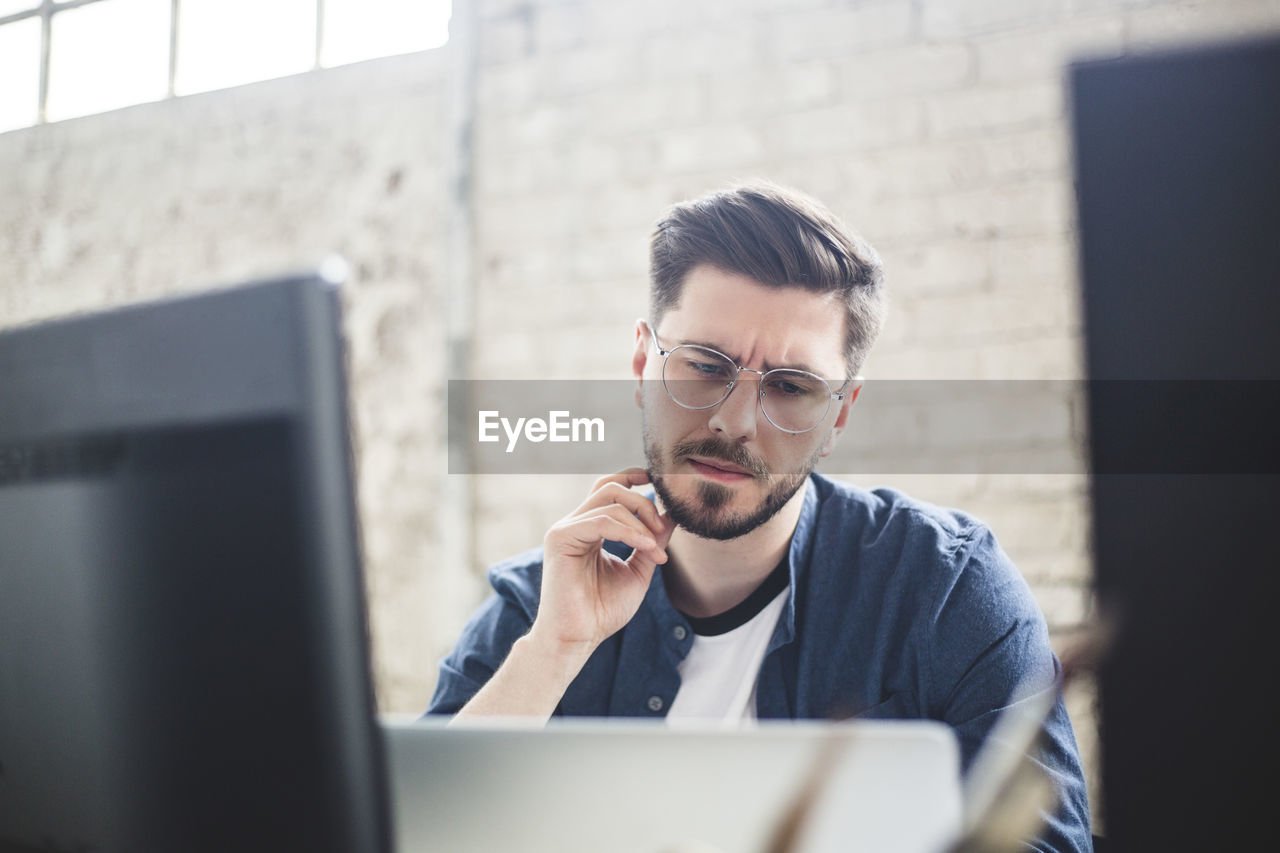 Thoughtful young it expert touching beard while looking at laptop in office