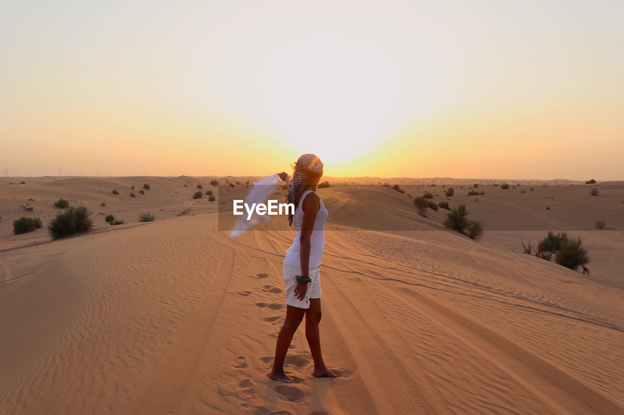 Side view of woman standing on sand at desert against sky during sunset