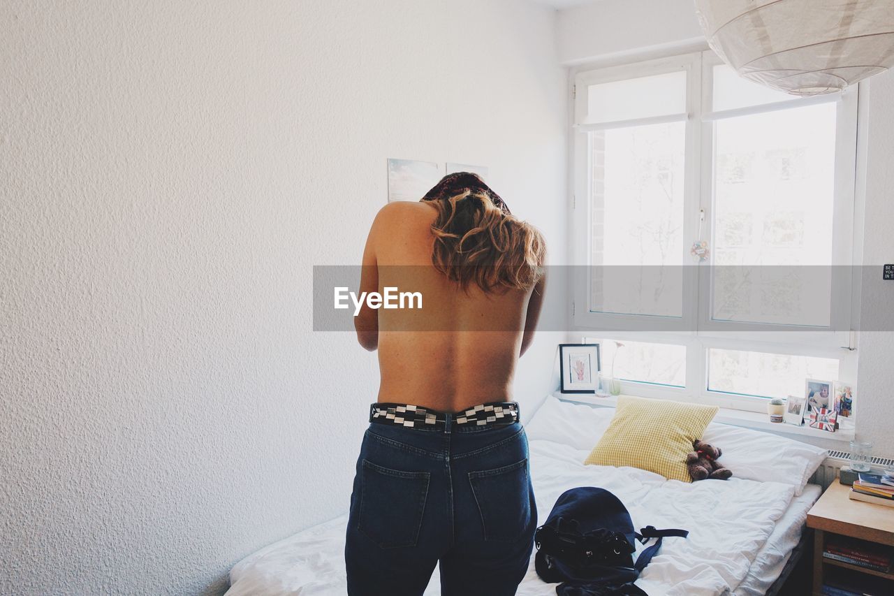 Rear view of shirtless young woman standing in bedroom