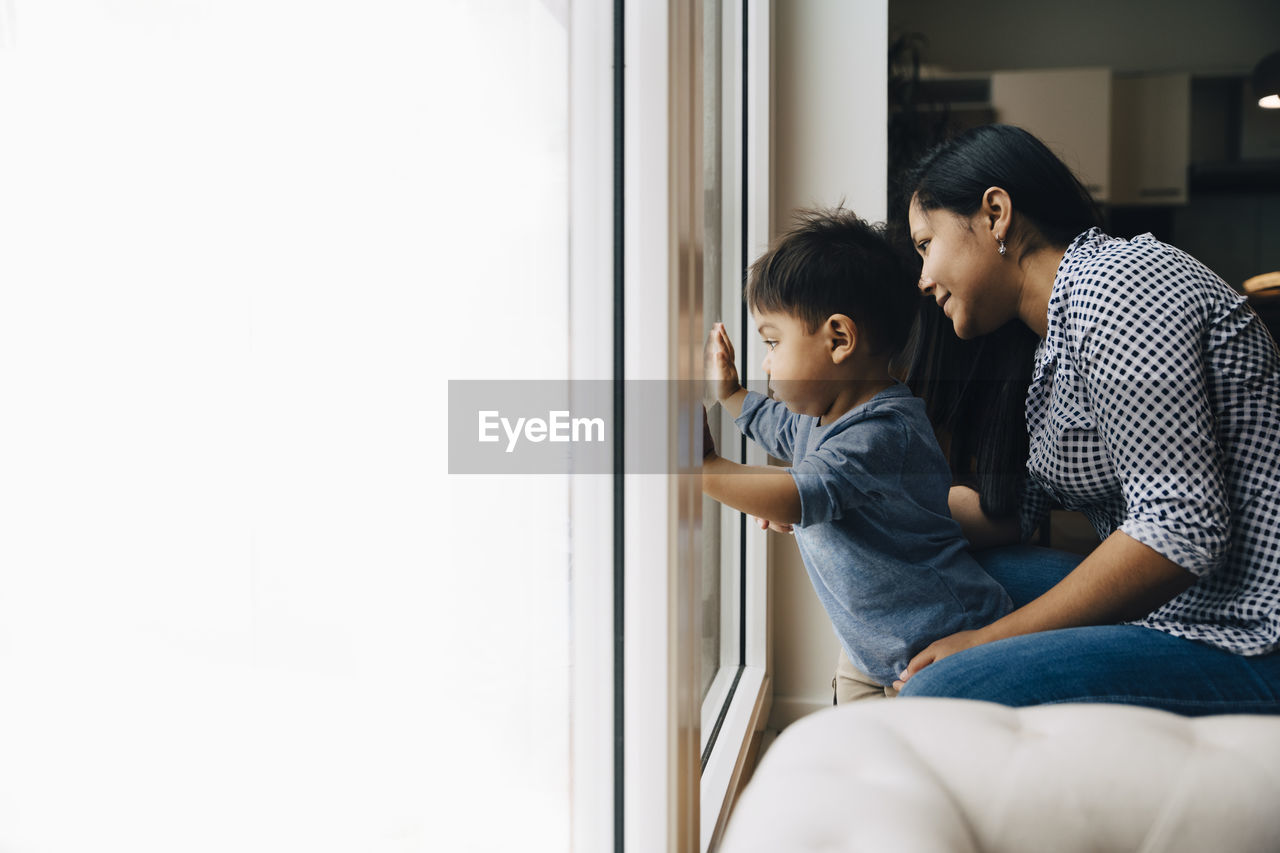 Side view of mother and son looking through window while sitting at home