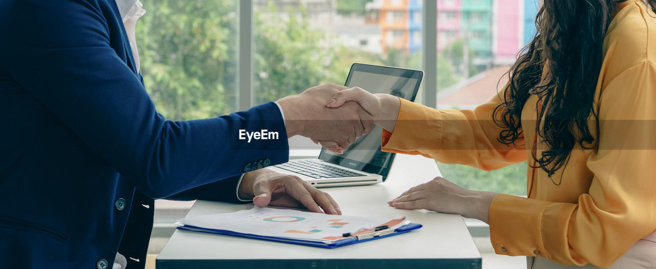 Two people shaking hands in front of a laptop. the laptop is open and has a picture of a person