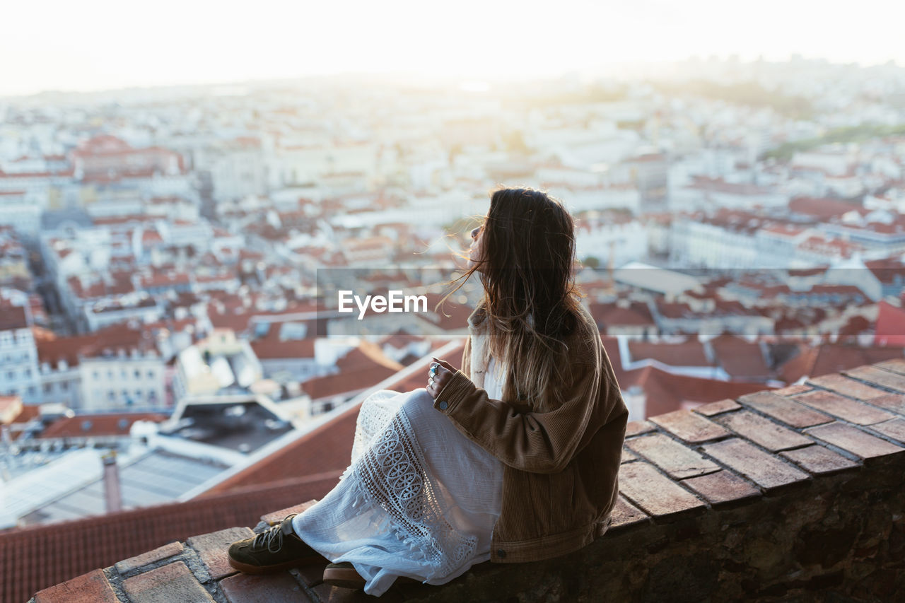 Side view of young lady in jacket and dress sitting on brick barrier and admiring cityscape in sunny morning in portugal
