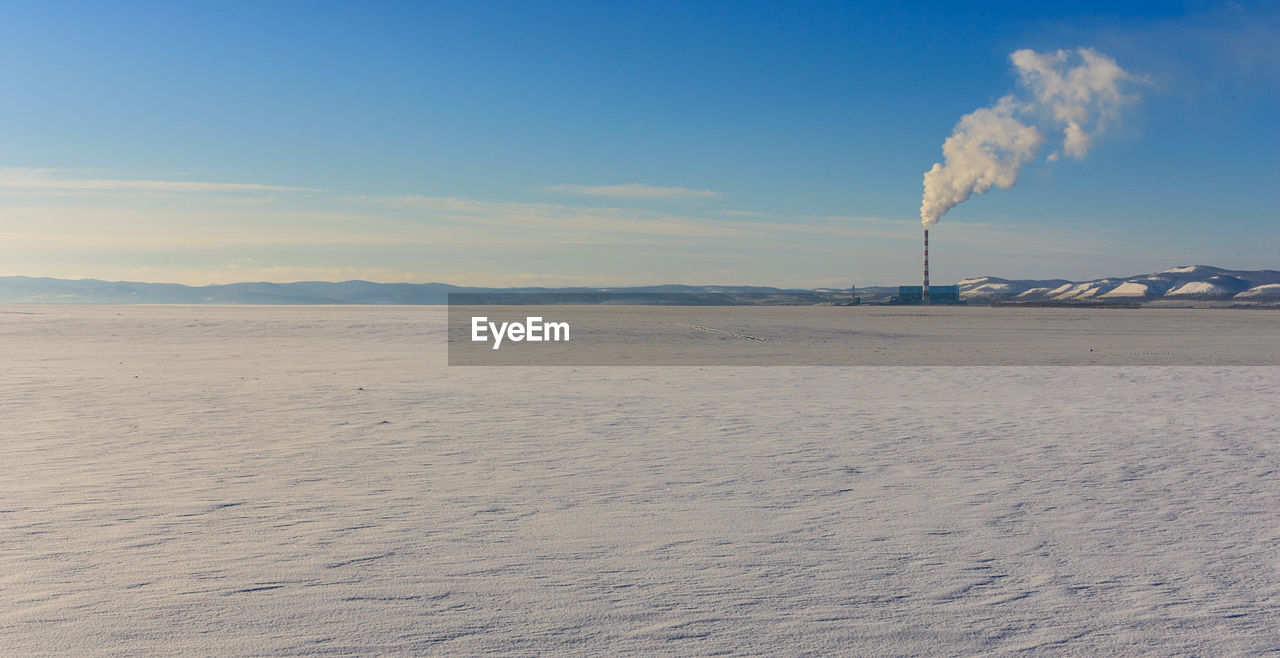 Distant view of smoke stack emitting pollution on snow covered field against sky