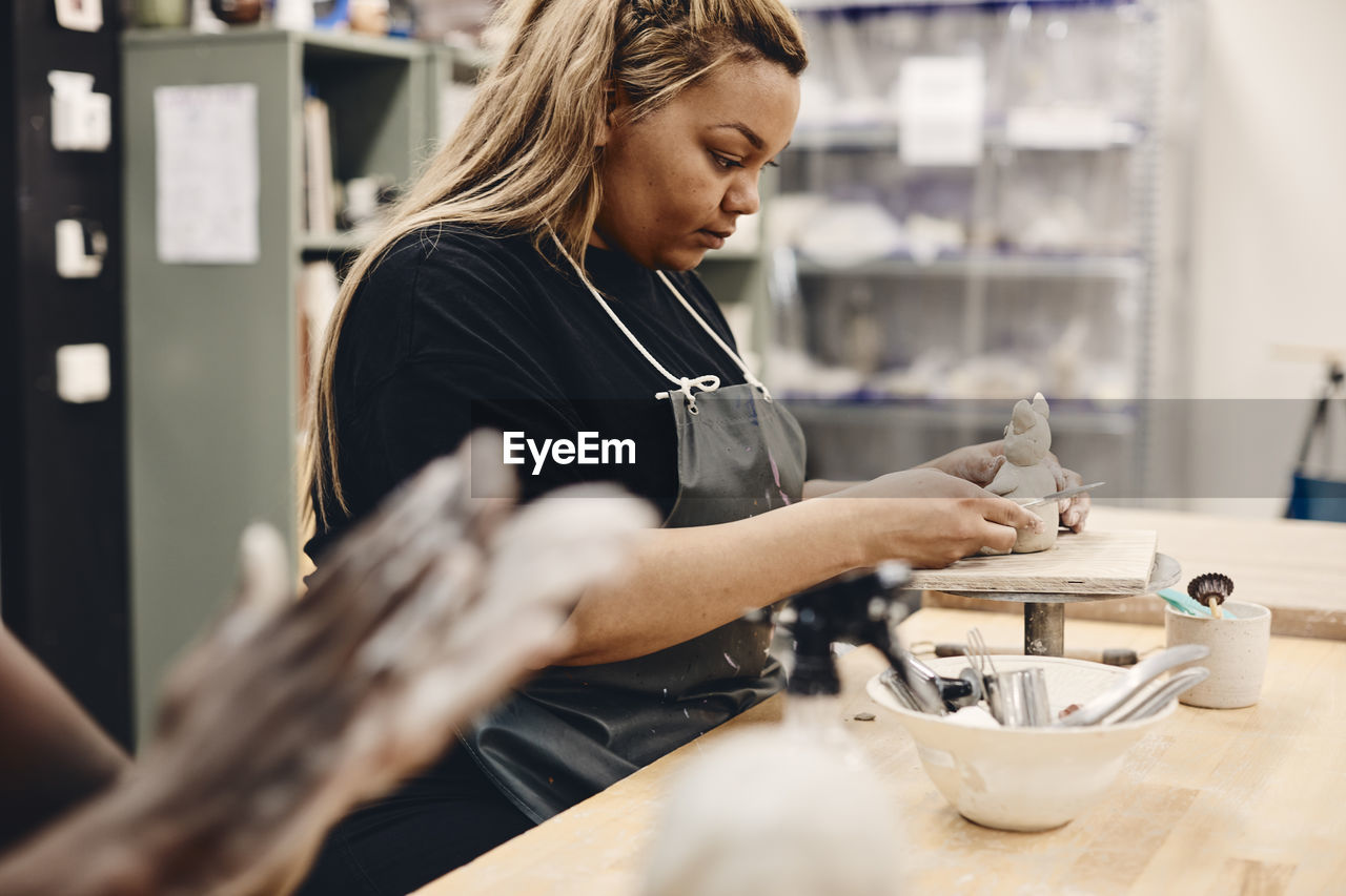 Young woman with work tool molding clay sitting at table in art class