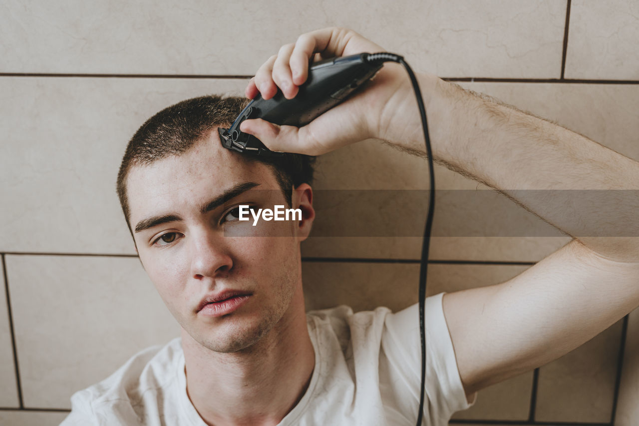 Lonely young man shaving hair with electric razor in bathroom