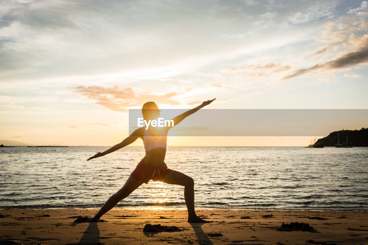 Silhouette young woman exercising at beach against sky during sunset