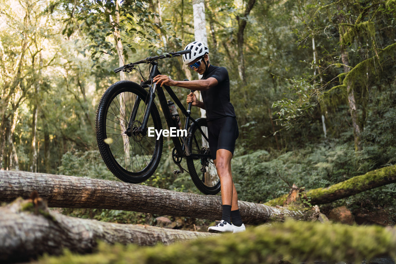 Low angle of fit sportsman in helmet and cycling goggles carrying bicycle on shoulder while walking on fallen tree trunk in woods