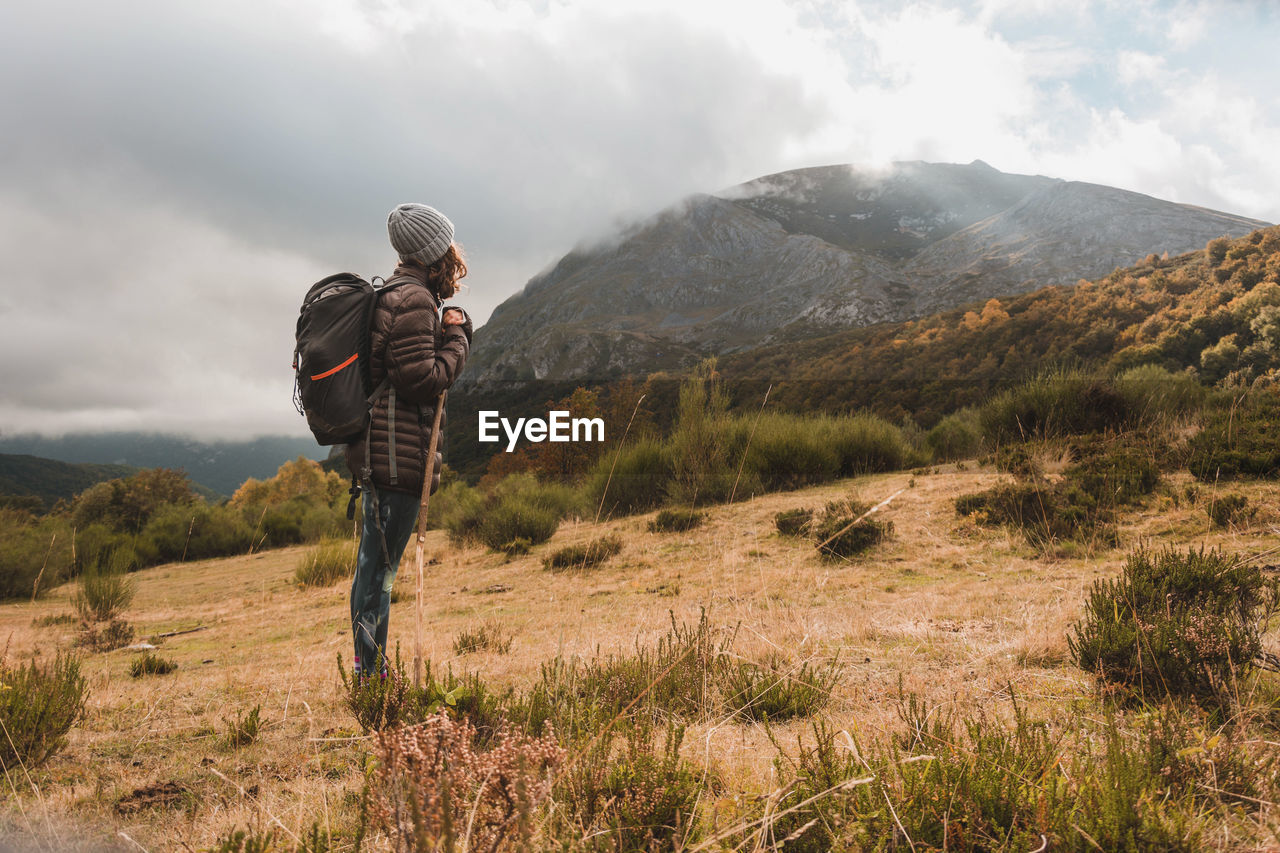 Side view of hiker with backpack standing on mountain against cloudy sky