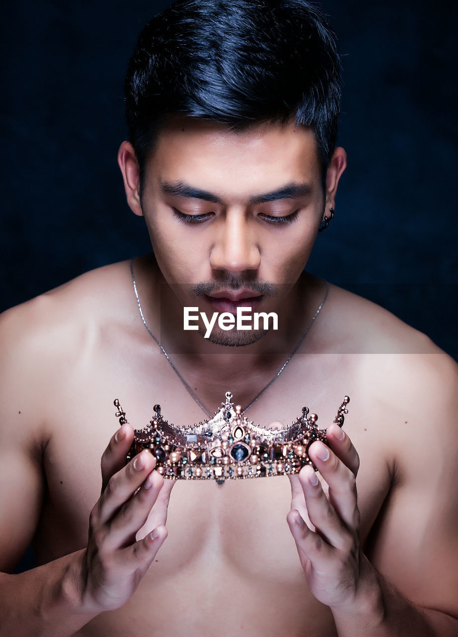 Shirtless young man holding crown against black background