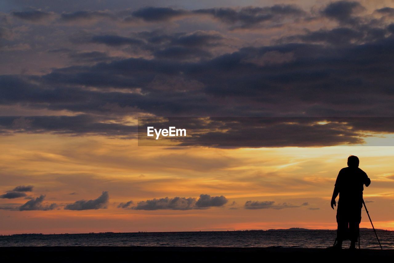 Rear view of silhouette man with tripod standing at beach during sunset