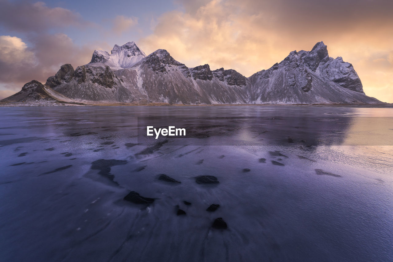 Spectacular nordic scenery of calm frozen lake near rocky vestrahorn mountain with snowy peaks during colorful sunset at stockness beach, iceland