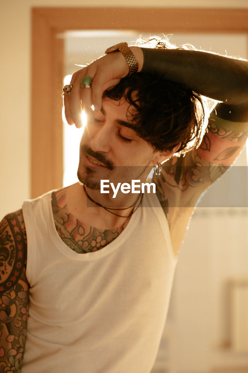 Young stylish guy with mustache and tattoos wearing white tank top and leaning against wall with closed eyes