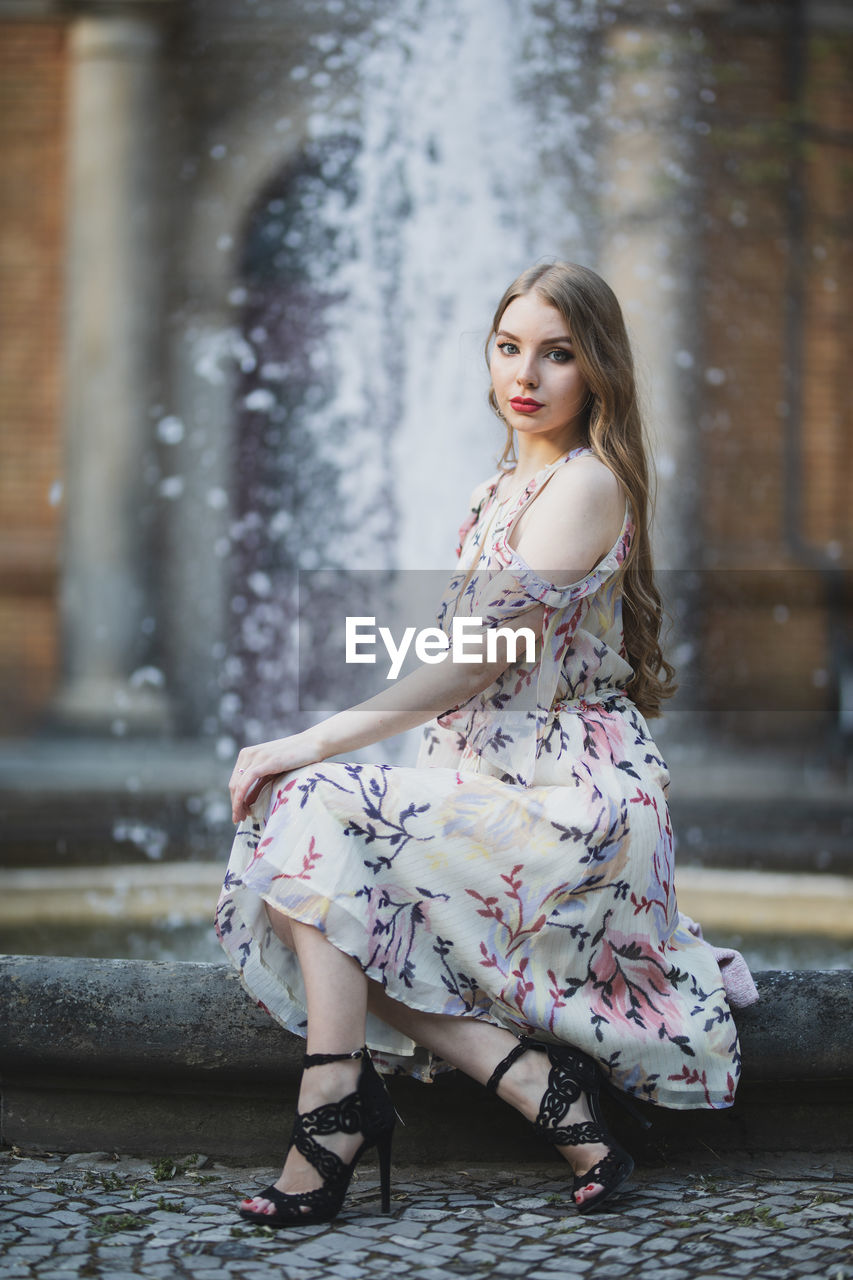 Portrait of beautiful young woman sitting against fountain in city