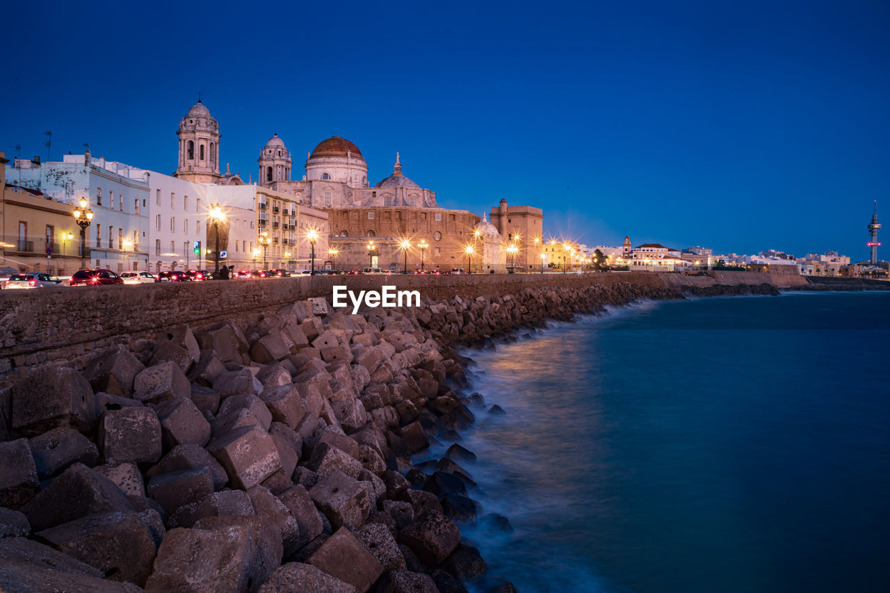 ILLUMINATED BUILDINGS AT WATERFRONT AGAINST BLUE SKY