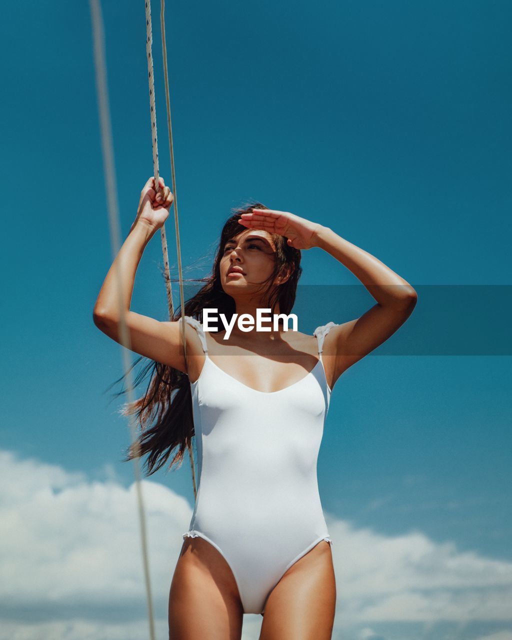 Woman in swimsuit shielding eyes while standing against blue sky